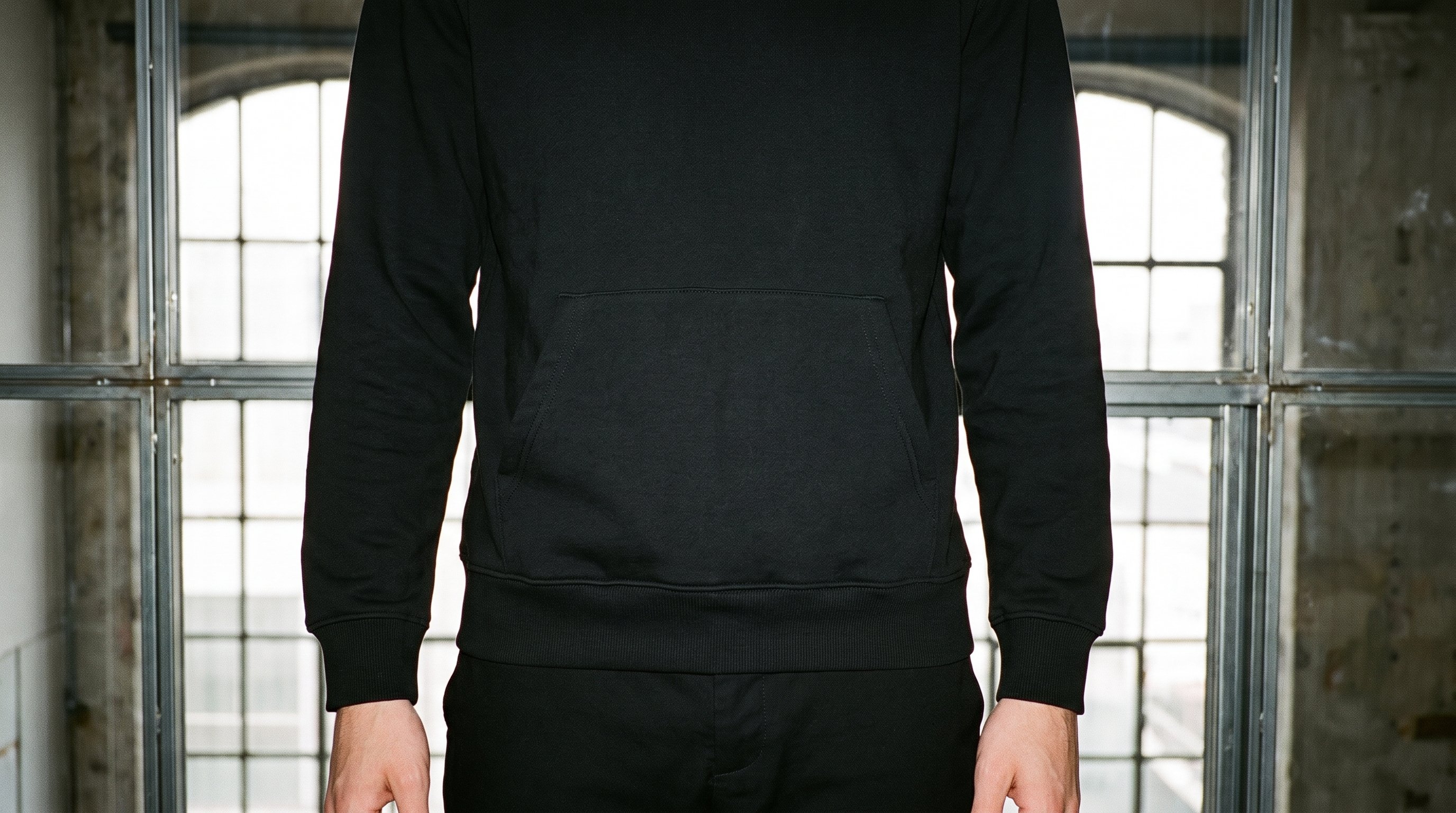 A male model is shown from the hips up in a front-facing, level pose wearing a black pullover hoodie. Massive loft windows form the golden backdrop.