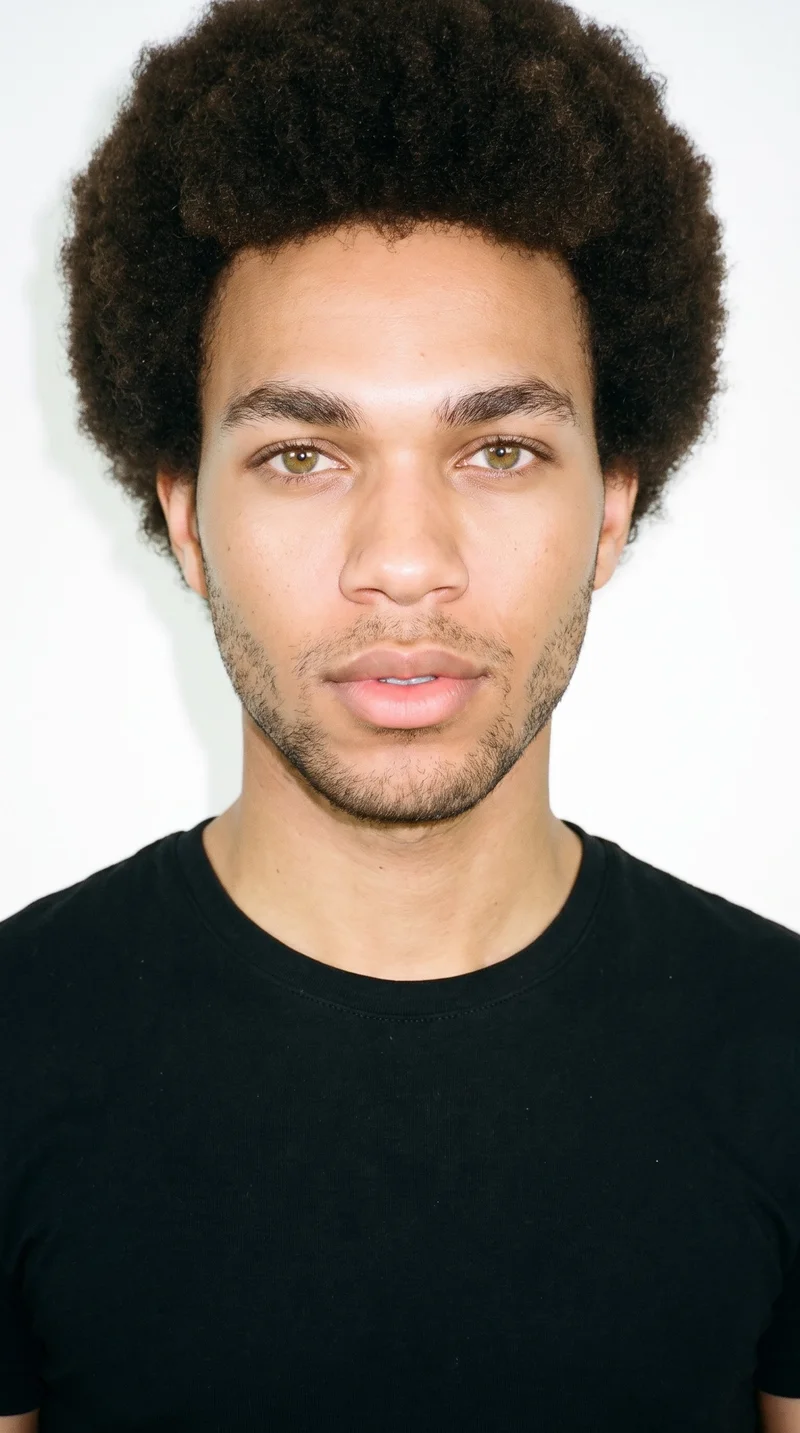 A 21-year-old African man with dark-brown skin and a warm golden undertone. His face has smooth, even, softly refined features in a balanced shape, with medium-thickness eyebrows, hazel eyes marked by green-gold flecks, and medium lips. His dark-brown hair is natural and tightly coiled in a full, rounded afro, and fine, closely trimmed stubble lightly covers the beard area.