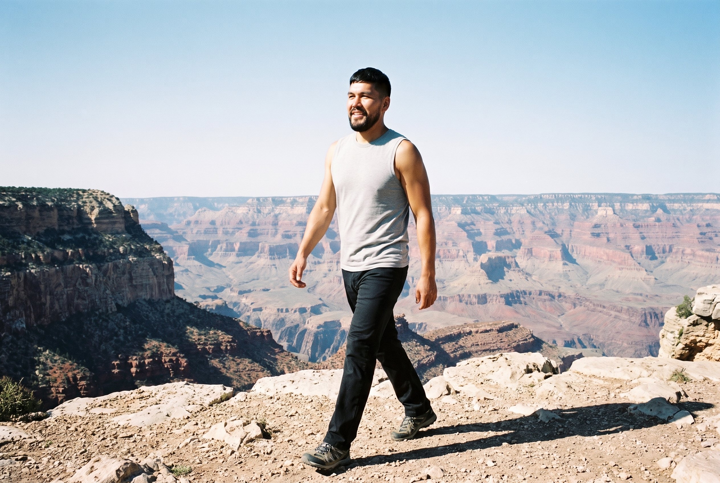 A 38-year-old Indigenous American man wearing a grey marl vest is shown full body from an angled front view while walking and smiling. The Grand Canyon Rim serves as the backdrop.