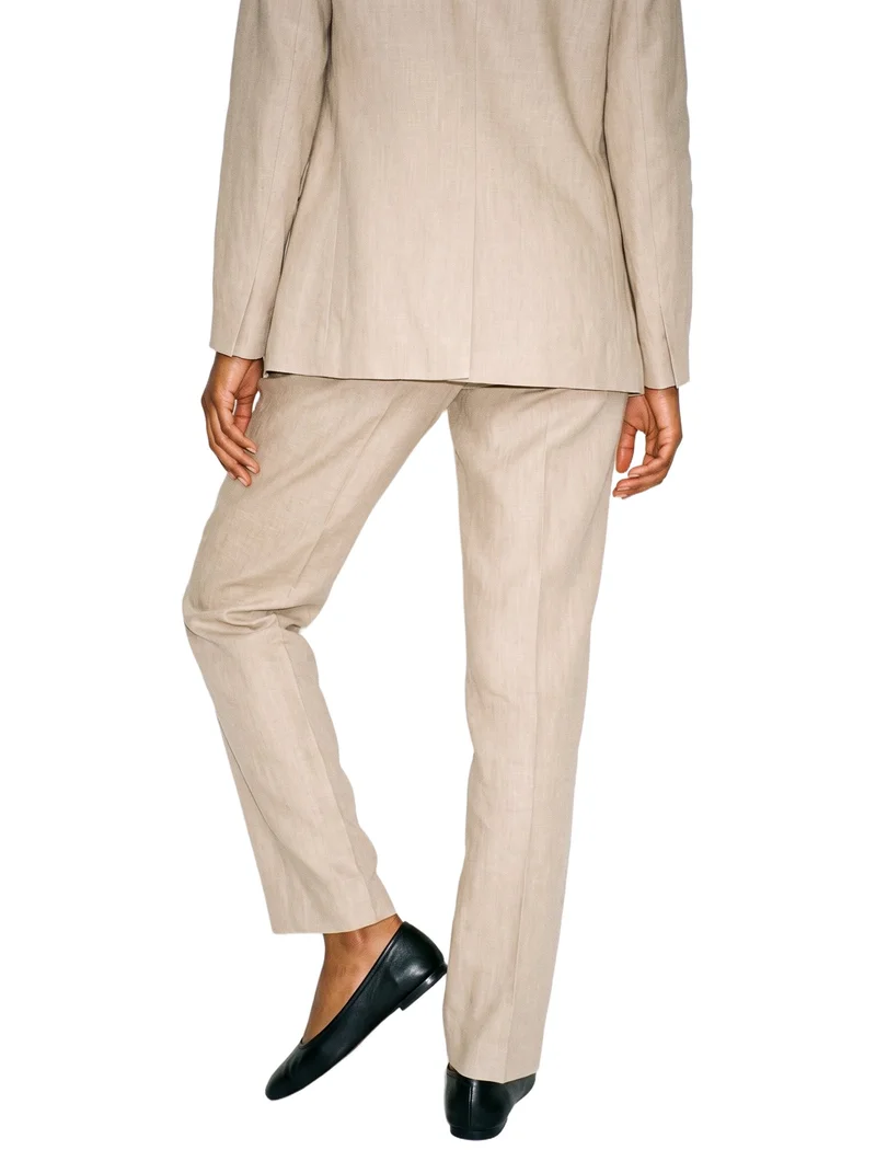 A lower-body rear view of a person wearing a beige linen trouser suit, posed with one heel lifted. The suit is shown against a pure white backdrop.