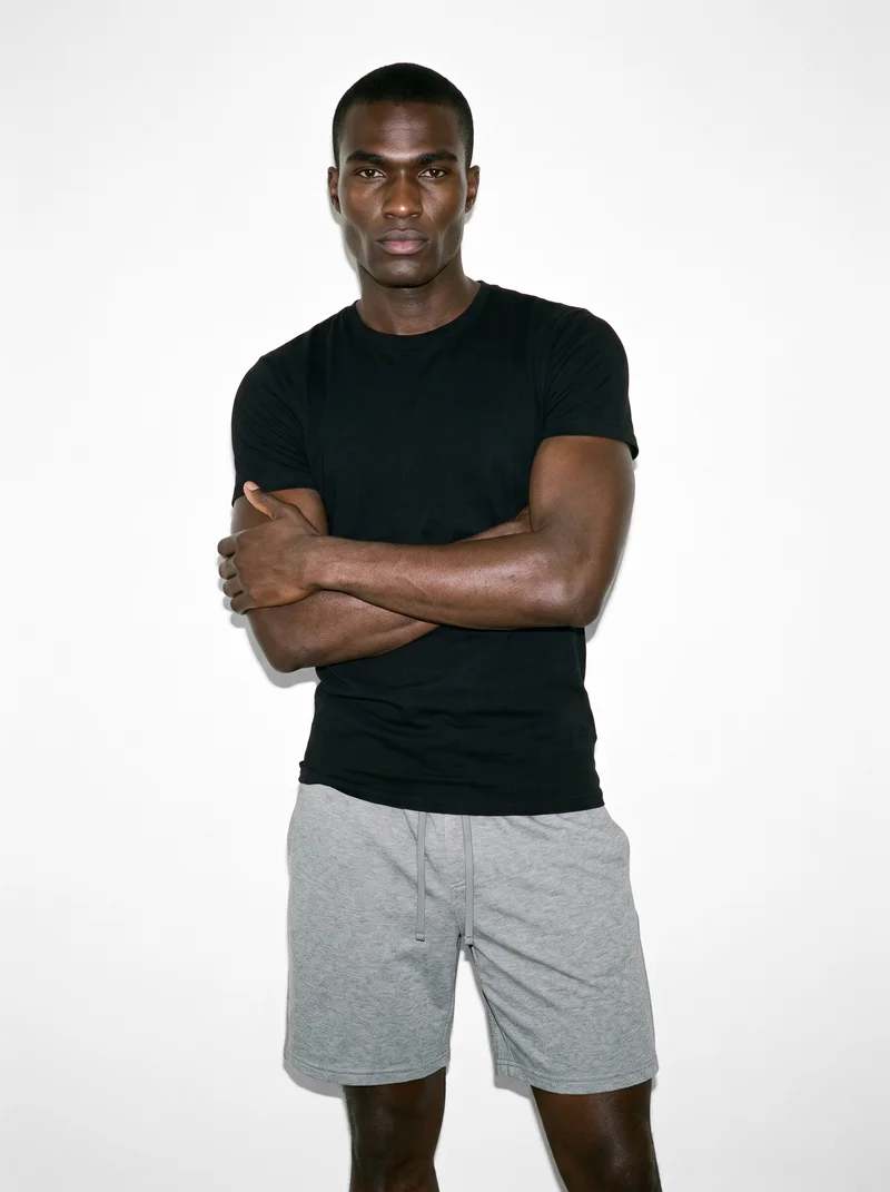 A 25-year-old African man wears grey gym shorts in a three-quarter-length front view. He stands in a self-hug pose with a neutral facial expression against a Seasalt studio-wall backdrop.