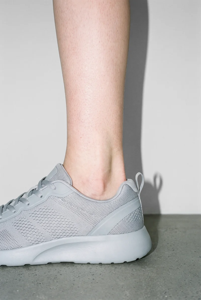 A grey mesh trainer from the women's range is shown in side view with the ankle flexed. The image is framed at the ankle against a Fog Grey studio-wall backdrop.