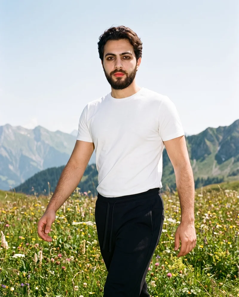A 22-year-old Arab man wears black joggers in a three-quarter-length photograph, shown from an angled front view while walking with a confident expression. A wildflower mountain meadow forms the backdrop.