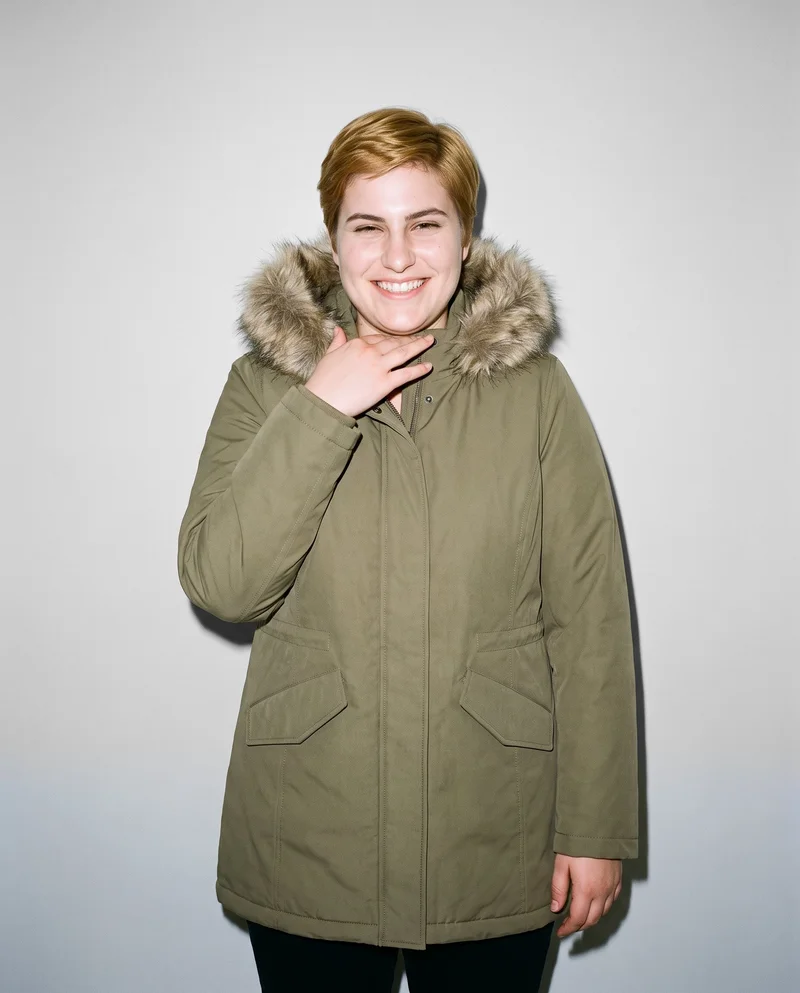 A 24-year-old Caucasian woman is shown from the upper body in a front view, wearing a women's khaki parka with a fur hood. She has one hand at her collarbone and a grin, against a pearl grey studio-wall backdrop.