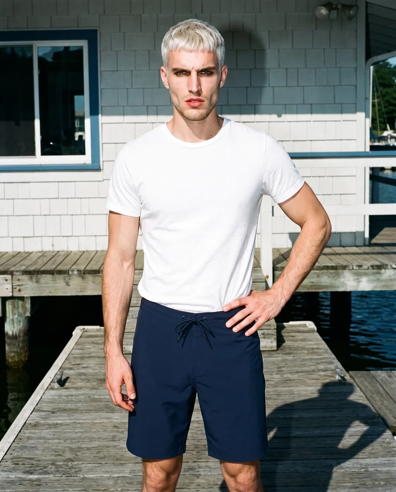 A 29-year-old Caucasian man wears navy board shorts in a front-facing three-quarter-length view, with one hand on his hip and an intense expression. A Yacht Club backdrop is visible behind him.