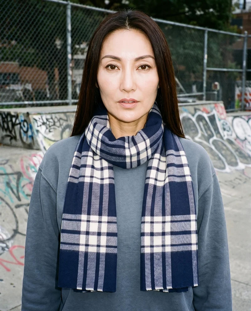 A 49-year-old Chinese woman is shown from the bust up in a front view, standing with her shoulders square and a neutral expression while wearing a women's navy checked wool scarf. A skate park forms the backdrop.