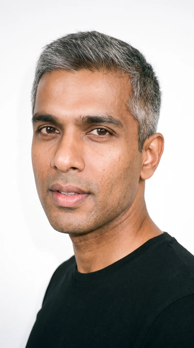 A 37-year-old Indian man with a South Asian appearance and copper-brown skin with an olive-green undertone. His square face has a broad forehead and a strong, defined jaw, creating an angular, structured appearance. He has medium-thick eyebrows, brown eyes, and very full lips. His gray hair is styled in a short, tapered crew cut that is slightly longer on top than at the sides, and he is clean-shaven with smooth skin across his face.
