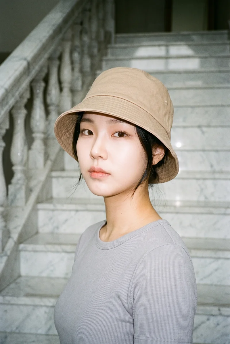 A 20-year-old Korean woman wears a women's beige bucket hat in a bust-length view from a front three-quarter angle. Her head is tilted with a confident expression, against a marble staircase backdrop.