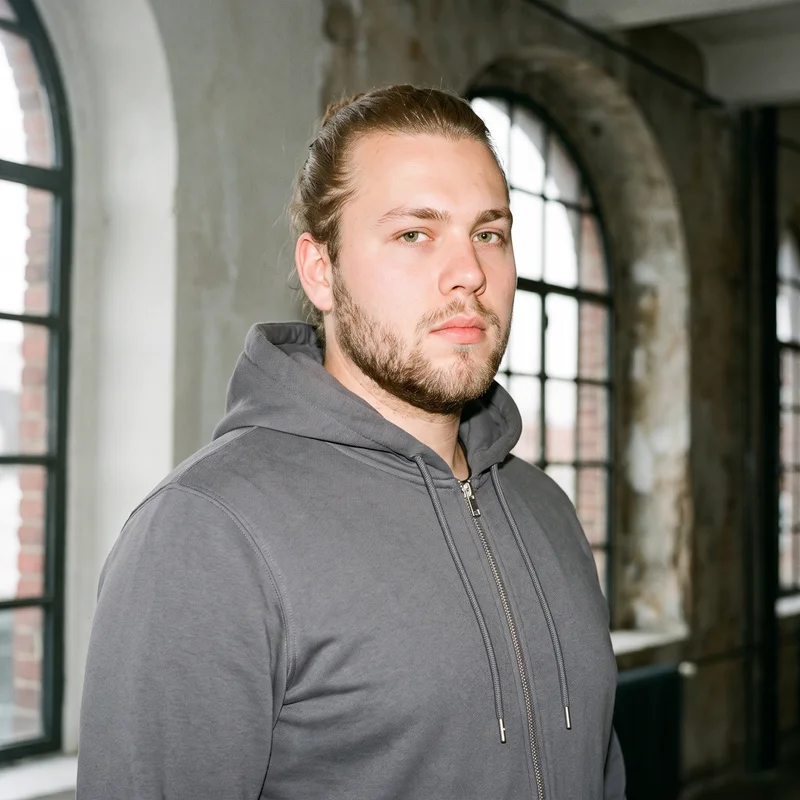 A 22-year-old Caucasian male model wears a charcoal cotton zip-up hoodie in a bust-length composition. He is shown from a front three-quarter angle with his shoulders square and a fierce facial expression, against a backdrop of large loft windows with golden light.