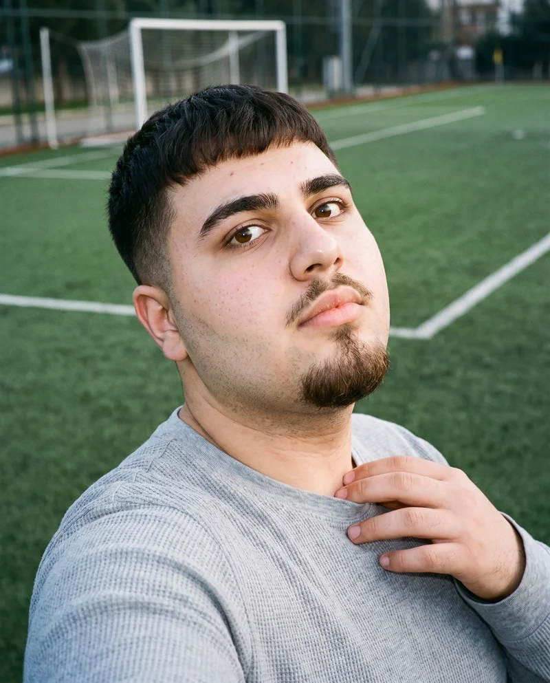 A 19-year-old Turkish man wears a men's grey thermal base layer in a front-view, over-the-shoulder selfie. The image has a fierce facial expression and a soccer pitch backdrop.