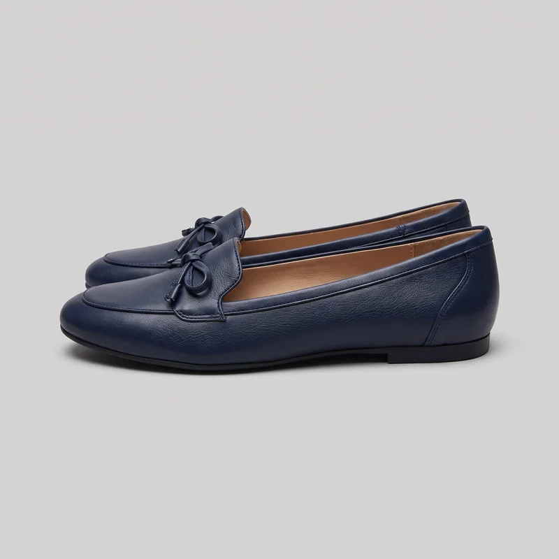 A pair of navy women's slip-on loafer flats is shown from a side three-quarter angle. The shoes have rounded almond toes, open toplines, pebbled uppers, apron-style vamp panels with raised piping and visible stitching, tied bow lace details with capped ends across the insteps, and flat soles with low stacked heels.