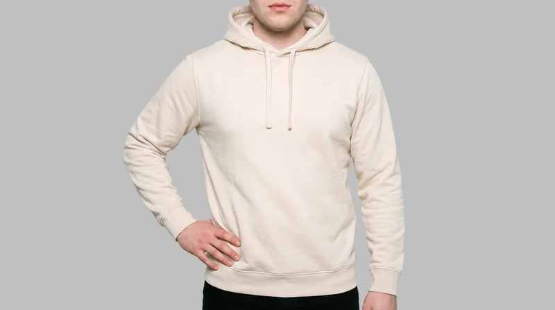 A male model is shown from the torso in a front view wearing a cream pullover hoodie, with one hand resting on his hip. The garment is presented against a flat silver mist backdrop.