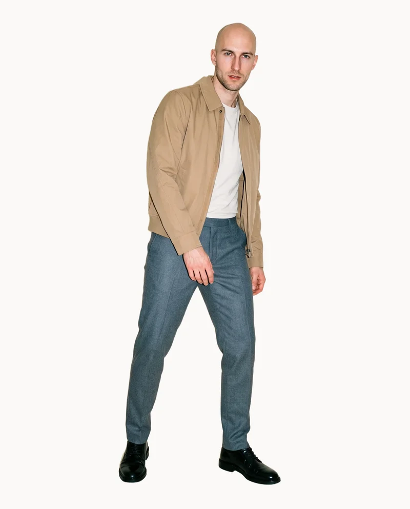 A 31-year-old Caucasian man wears a men's khaki bomber jacket in a full-body front view, posed with a waist twist and a curious facial expression. A Chantilly Lace flat-color backdrop frames the figure.