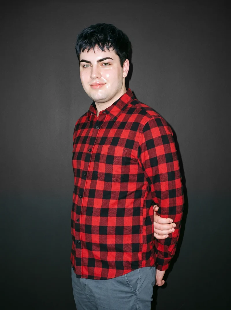 An 18-year-old Caucasian man wears a red check flannel shirt in a three-quarter-length composition. He is shown from a front three-quarter angle with one elbow positioned behind his back and a smile, against a carbon studio-wall backdrop.
