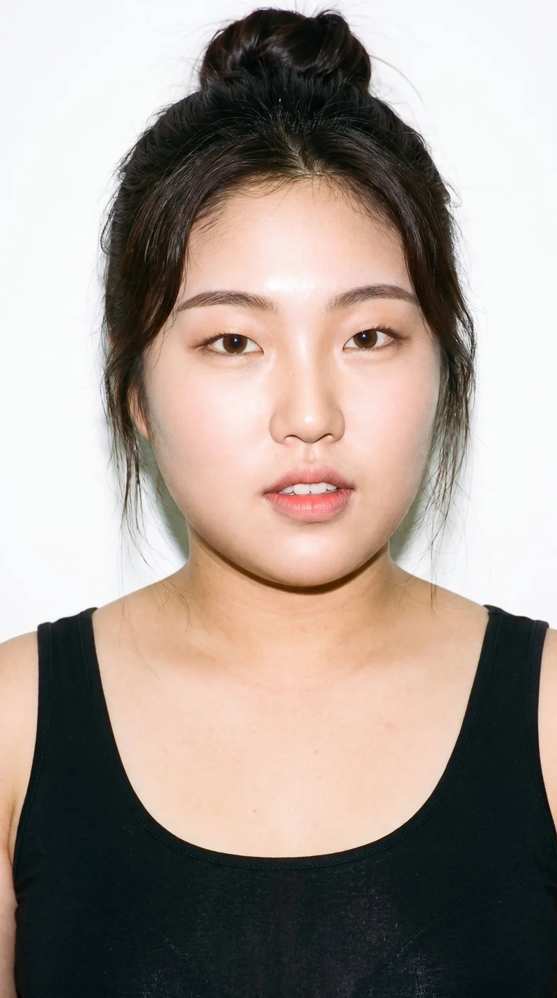 An 18-year-old Korean woman with medium skin and a cool undertone has a softly rounded, doll-like face with large, even features. She has thin eyebrows, full lips, and very dark, near-black eyes. Her dark-brown hair is gathered into a loose, messy bun with softly undone strands.