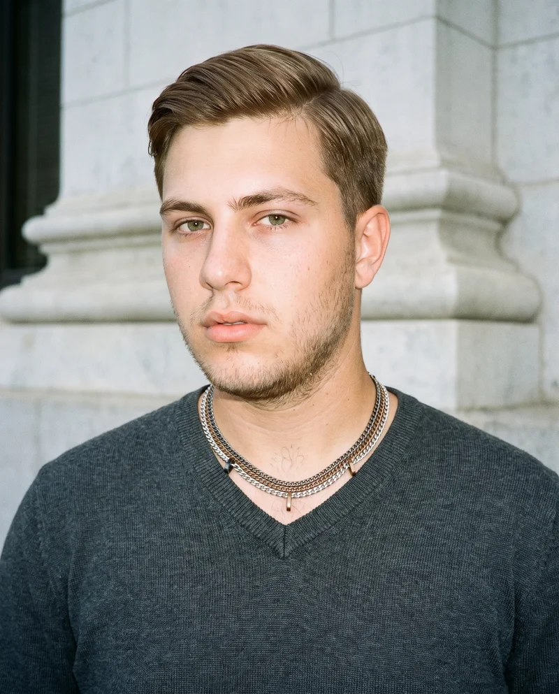 A 19-year-old Caucasian man is shown from the bust in a front view with one shoulder forward and a soft expression, wearing a men's mixed-metal layered necklace. Courthouse steps form the backdrop.