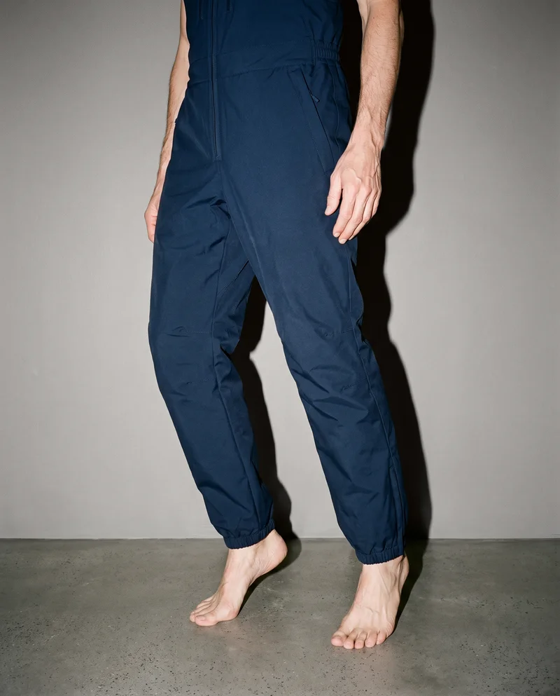 Men's navy insulated salopettes shown on a model from a front three-quarter angle, framed on the lower body while standing on tiptoes. A studio grey backdrop is visible.