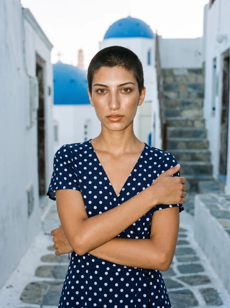 A 21-year-old Turkish woman wears a polka-dot wrap dress in an upper-body front view with her arms crossed and a sultry expression. White steps form the backdrop.