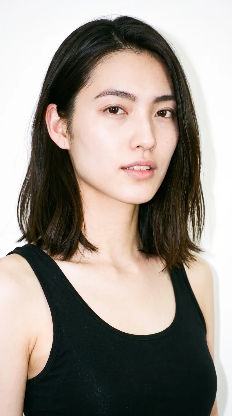 This 24-year-old Japanese woman has light porcelain skin with a cool undertone and East Asian facial features. Her face is oval, with balanced proportions and a softly rounded jaw. She has thick eyebrows, very dark near-black eyes, and full lips. Her dark-brown hair is styled in a long bob that falls between the chin and shoulders.