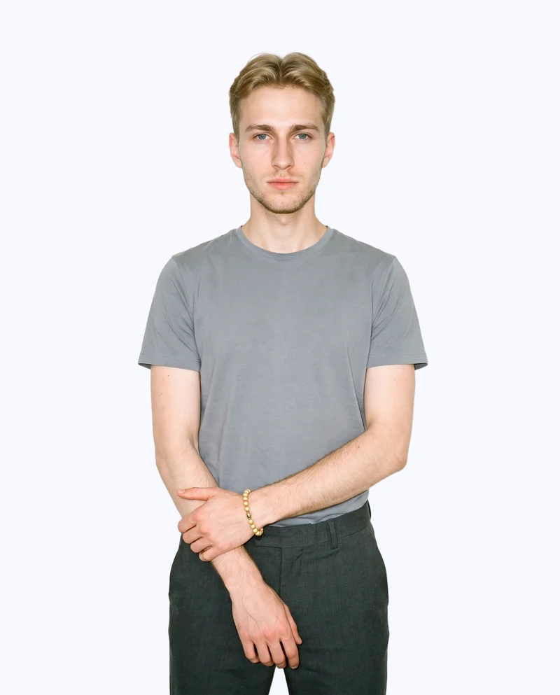 A 21-year-old Slavic man is shown in an upper-body front view with his forearms folded, wearing a men's gold beaded bracelet. The image has a serious expression and a flat porcelain backdrop.
