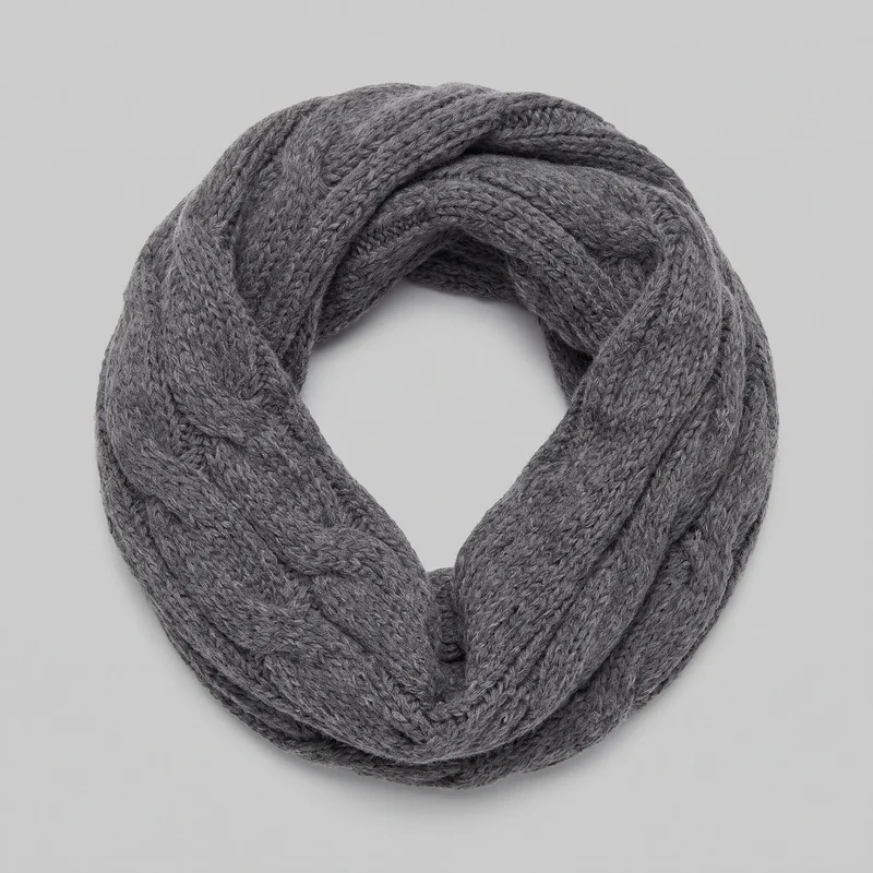 A complete women's grey knit snood is shown as a coiled, closed loop on a plain surface. Its rounded tubular form features raised cable-knit bands, braided ribbed sections, and open stitch channels, with softly rolled edges and no visible closure or added embellishments.
