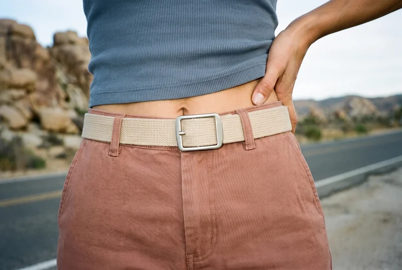 A woman's beige canvas belt is shown around the waist in a front-view waistline crop, with the model twisting at the waist. A Monument Valley Road backdrop is visible.