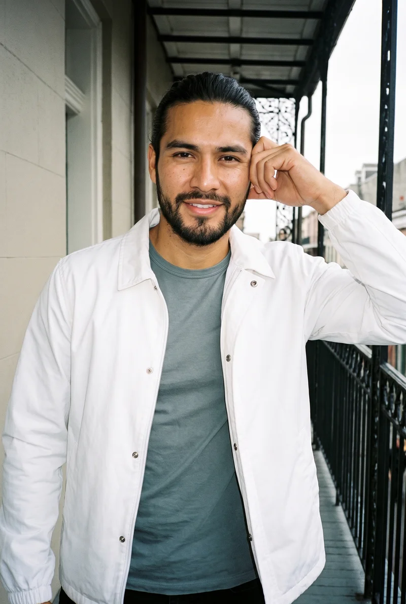 A 27-year-old Hispanic man is shown from the upper body in a front view, wearing a white windbreaker and smiling with one hand at his temple. A New Orleans balcony serves as the backdrop.