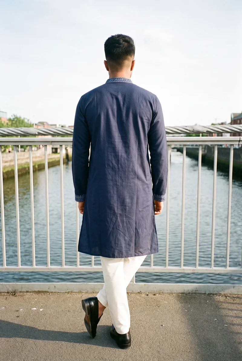 A 26-year-old Indian man wears a navy embroidered kurta in a full-body rear view, standing with his ankles crossed. A bridge pedestrian backdrop is visible behind him.