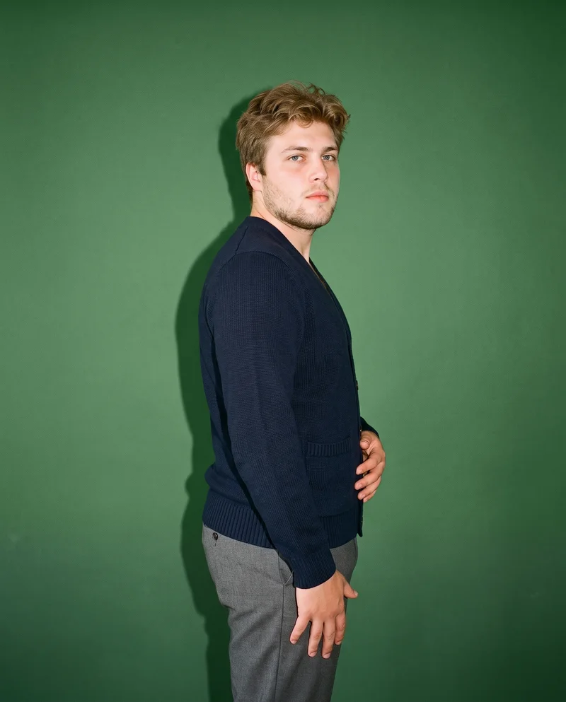 An 18-year-old Caucasian male model wears a men's navy wool cardigan in a three-quarter-length side view, posing with one hip out and an intense facial expression. The hunter green studio-wall backdrop frames the composition.