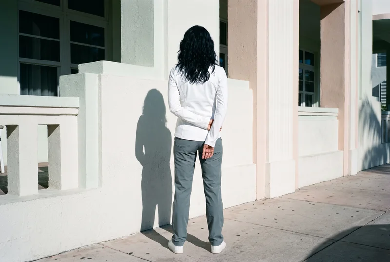 A 45-year-old Hispanic woman wears a white women's track jacket in an environmental back view with her arms low. A Miami Beach Deco Hotel serves as the backdrop.