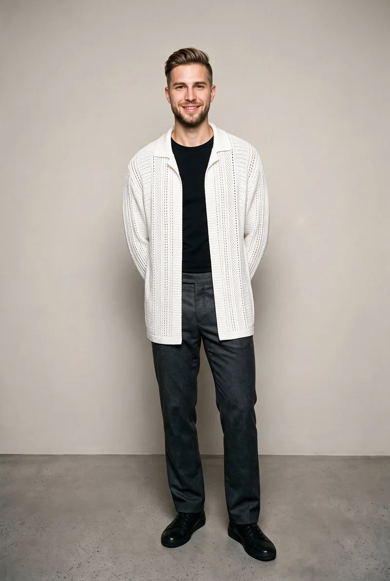A 22-year-old Caucasian man wears a white crochet cover-up in a full-body front view, standing with his hands behind his back and smiling. A greige studio-wall backdrop frames the composition.