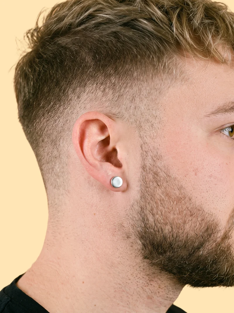 A man's ear shown in side view with a silver stud earring and the ear angled forward. The image uses a flat peach backdrop.