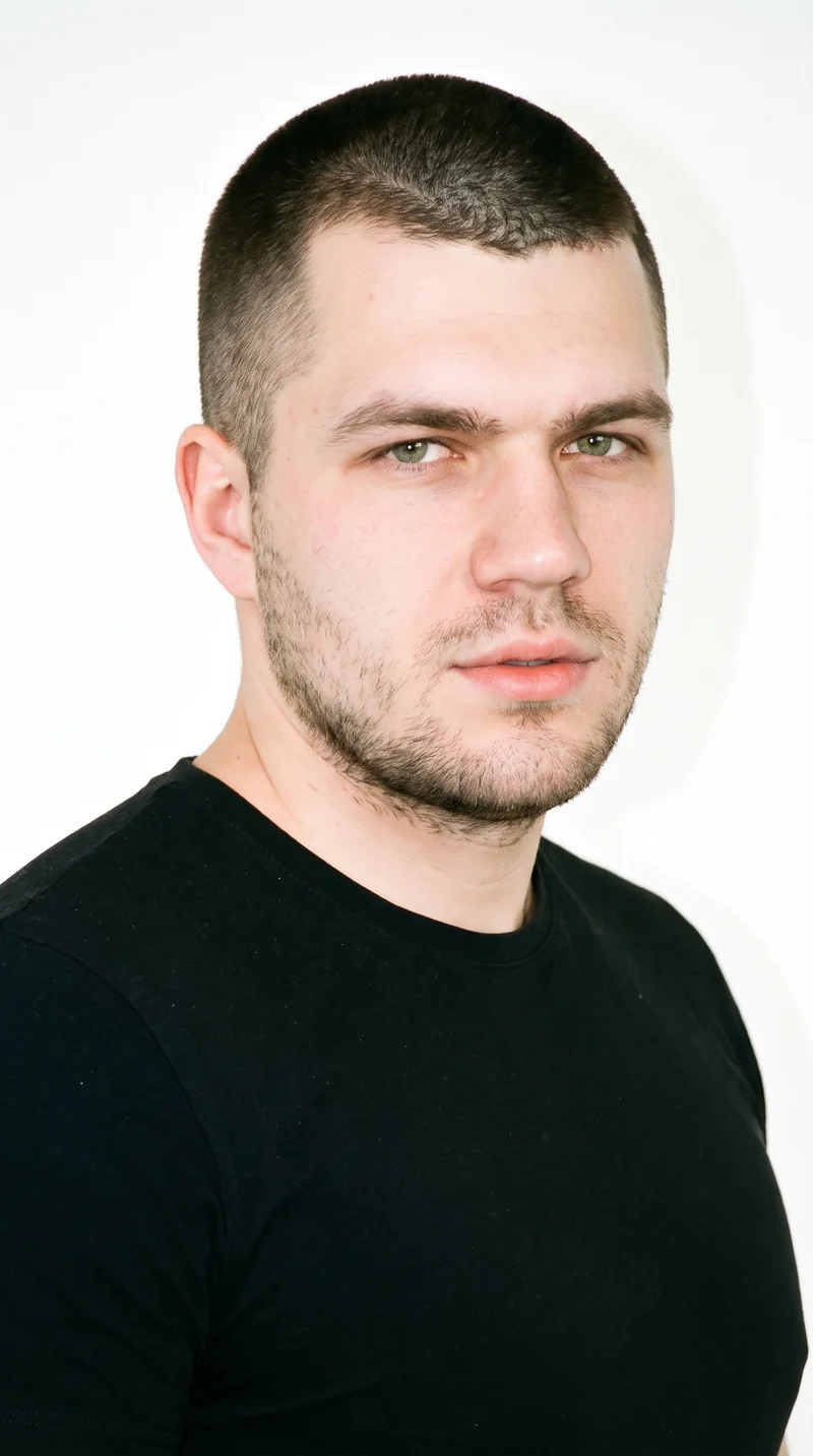 A 23-year-old Slavic man with a light porcelain complexion and cool undertone has a rugged, weathered face with characterful features and a heavy jaw. His eyebrows are thin, and his eyes are green. He has medium lips and dense, heavy stubble forming a dark, defined layer across the beard area. His black hair is clipped very short and evenly in a buzz cut.