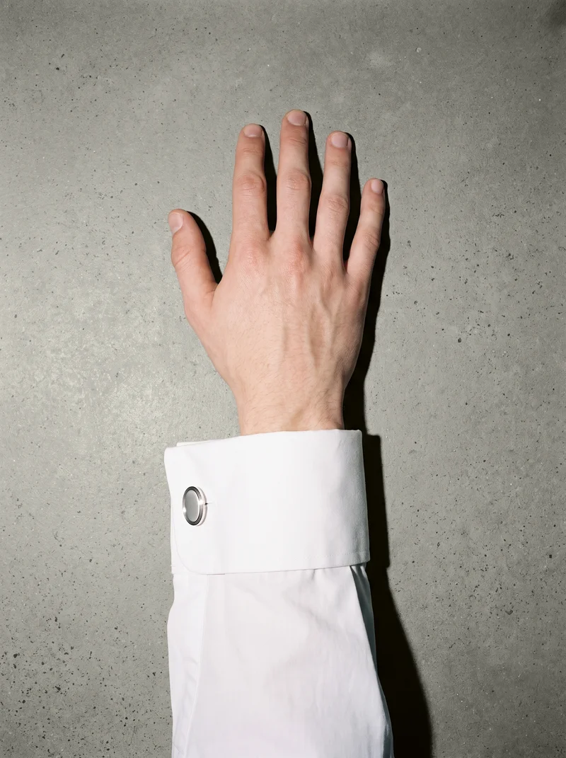 A hand and wrist are shown from a top view with the hand held flat, displaying silver classic cufflinks. An alabaster studio-wall backdrop surrounds the composition.