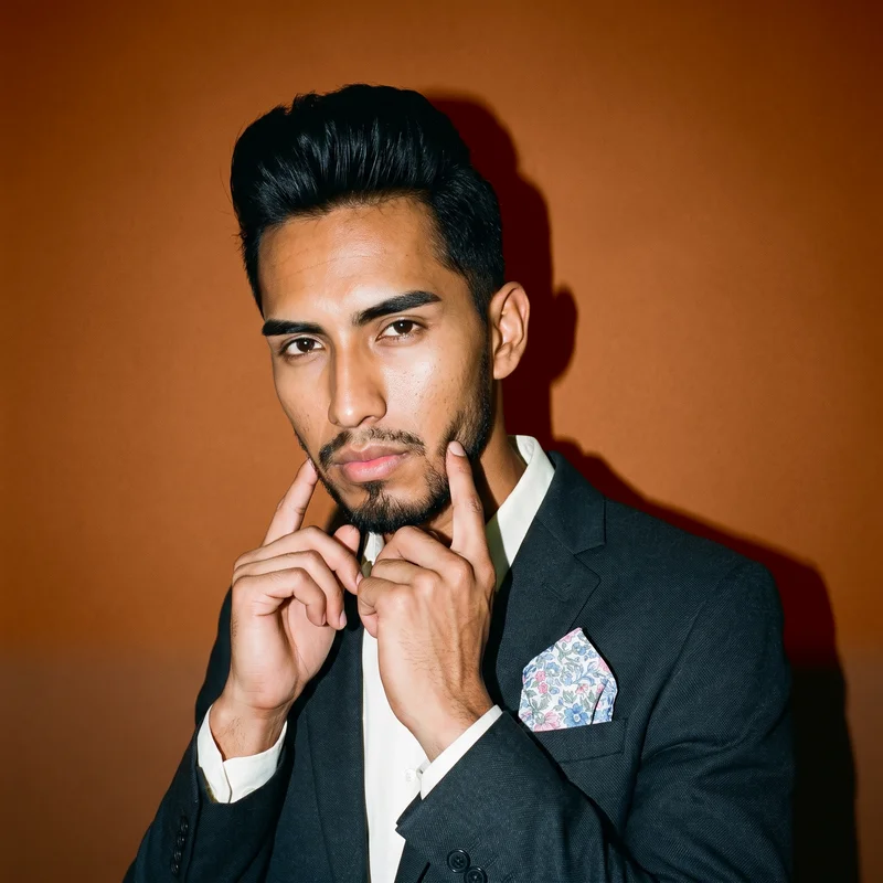 A 20-year-old Hispanic man is shown in a bust view from a front three-quarter angle, wearing a floral print pocket square with his hands at his jaw and a confident expression. A saddle brown studio-wall backdrop fills the background.