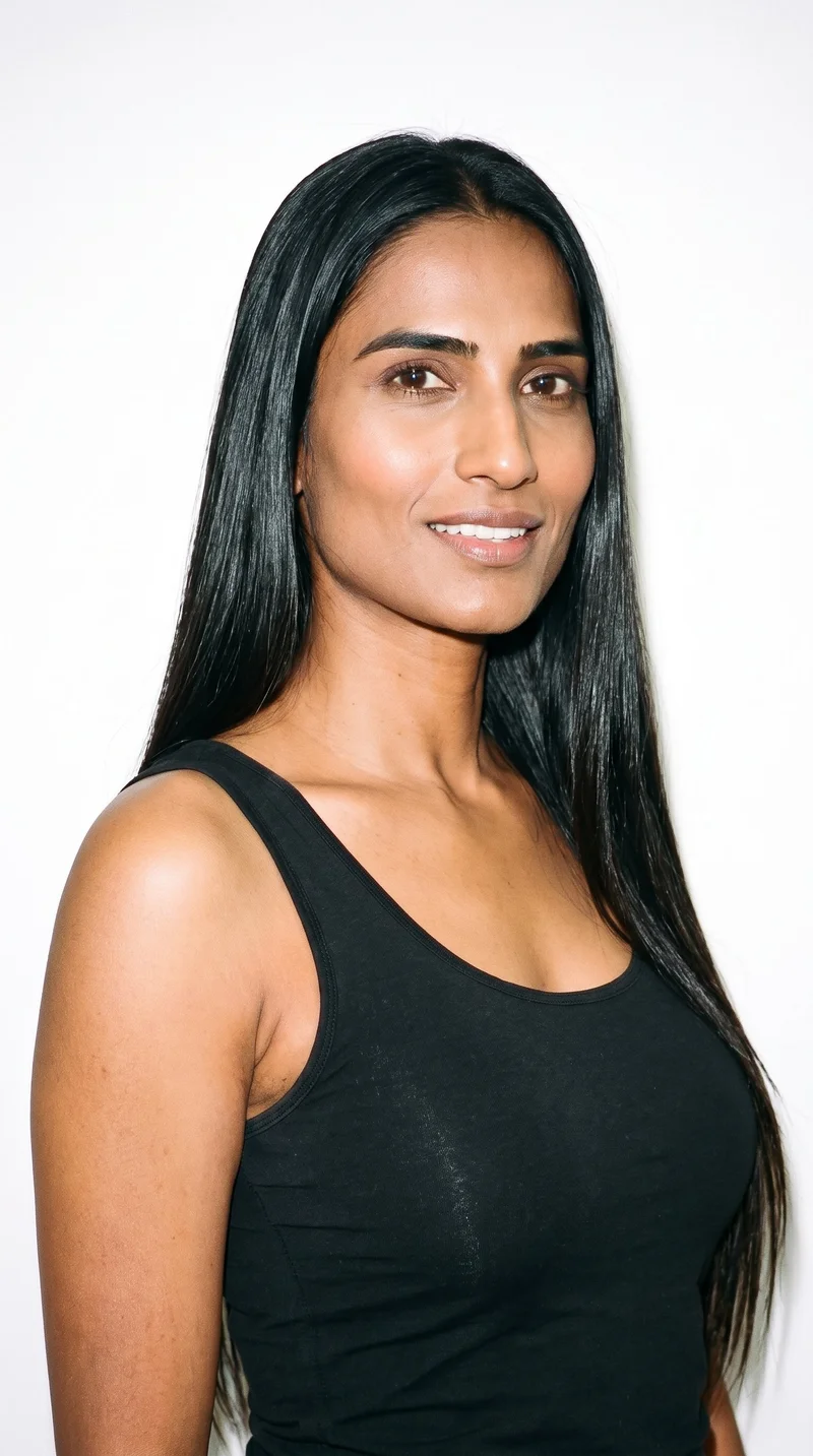 A 37-year-old Indian woman with medium-brown caramel skin and a warm olive-green undertone. Her triangular face has a broad jaw that narrows toward the forehead, emphasizing the lower face. She has thick eyebrows, very dark near-black eyes, and full lips. Her deep black hair is long and straight, falling without waves or layers.