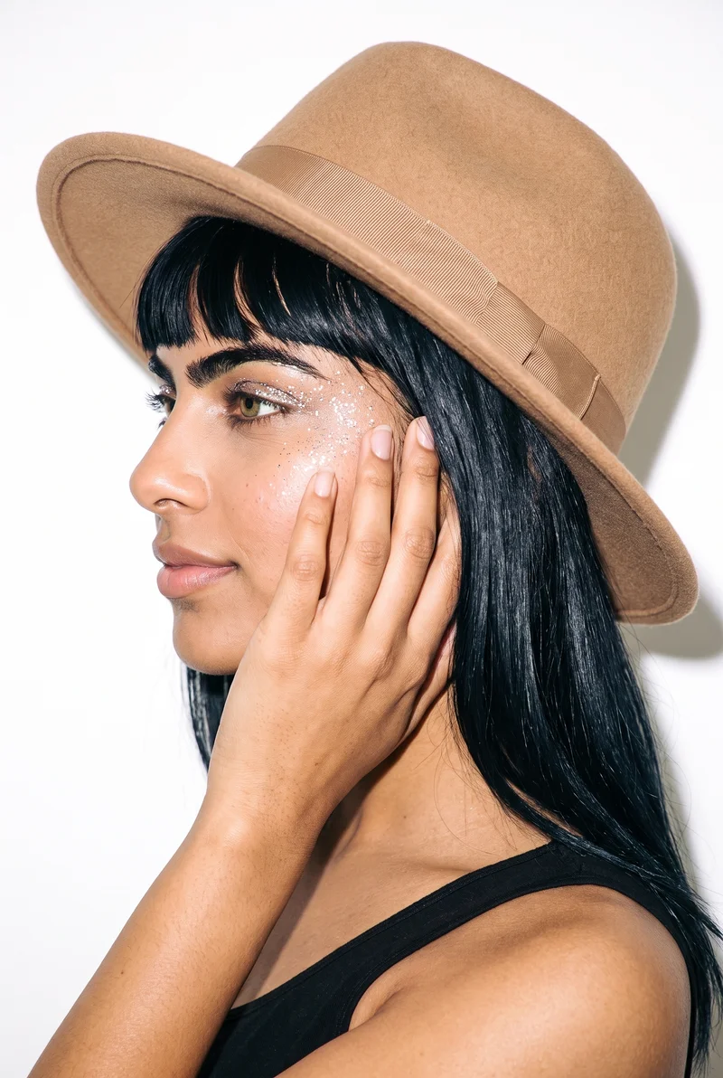 A 26-year-old Indian woman wears a women's camel felt fedora in a head-framed side view, with one hand at her face and a smiling expression. An Amsterdam canal house serves as the backdrop.