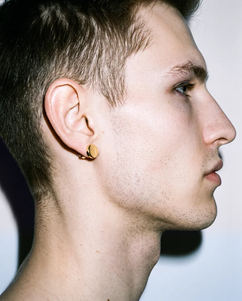 A side view of a male ear from the back, showing a gold stud earring against a porcelain backdrop.