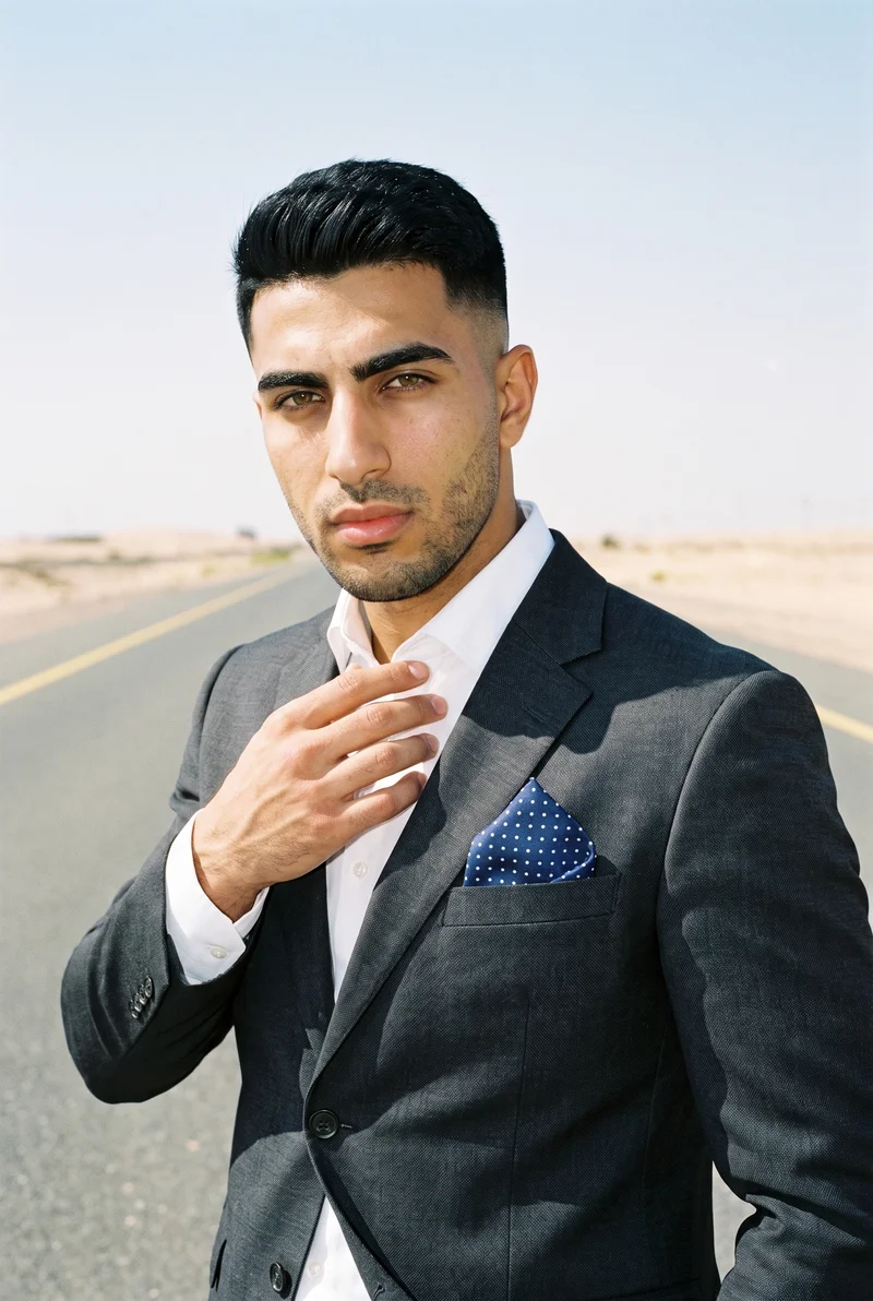 A 25-year-old Arab man is shown from the upper body in a front view, with his fingers at his chest and an intense expression. He wears a polka-dot pocket square, with an endless desert highway backdrop.