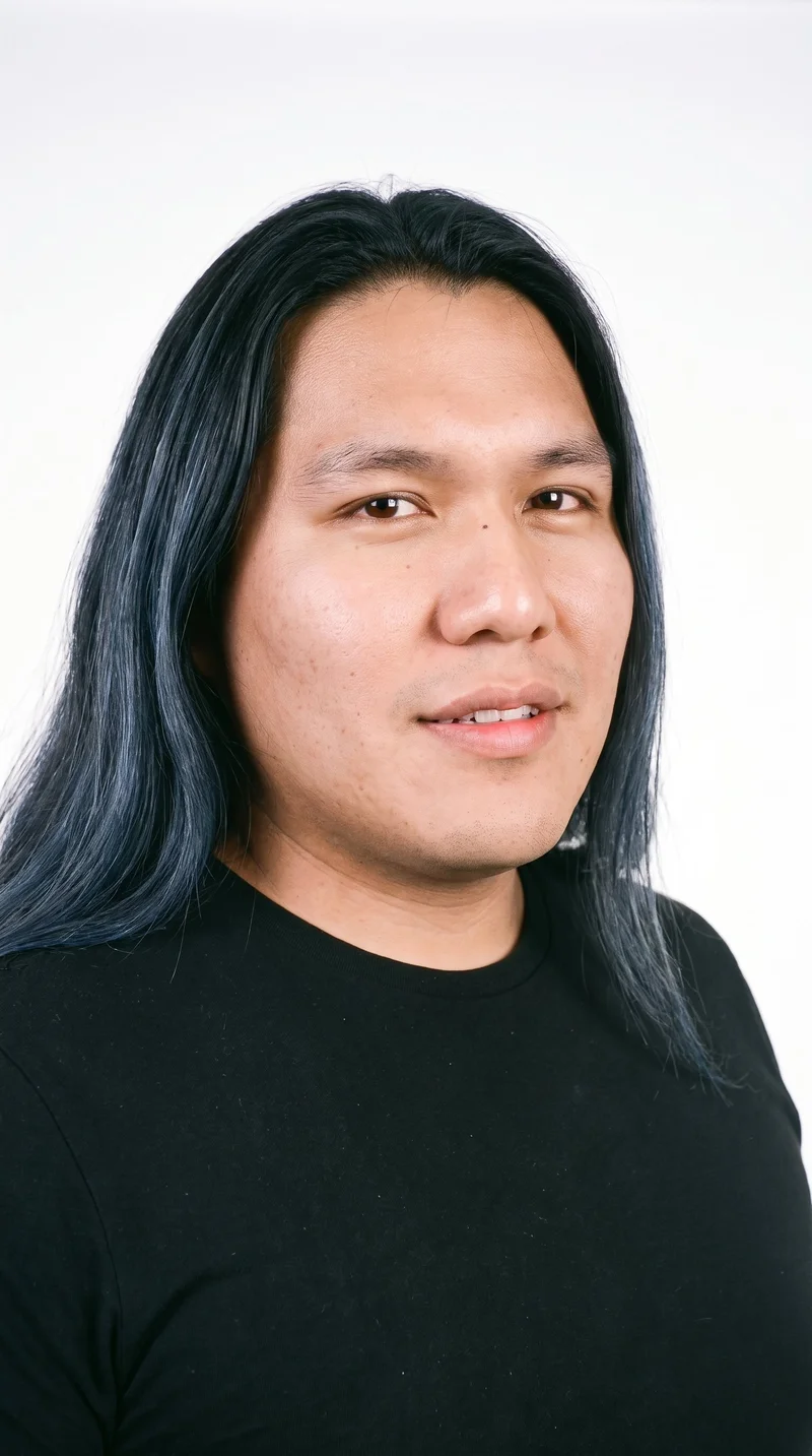 A 28-year-old Indigenous American man with a medium, evenly warm skin tone and a red-copper undertone. His oval face has balanced proportions and a softly rounded jaw, with medium-thickness eyebrows, brown eyes, and full lips. His deep black hair is long, worn loose past the shoulders with natural volume, and his face is clean-shaven.