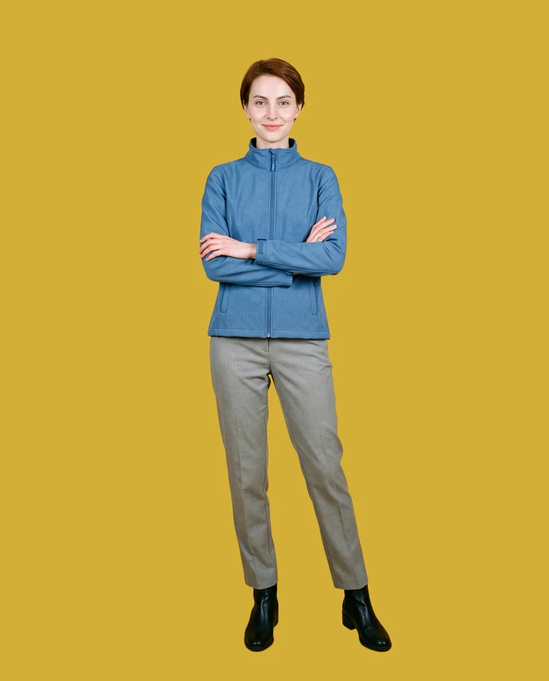 A 24-year-old Slavic woman wears a women's blue softshell jacket in a full-body front view, standing with her arms lowered and smiling. The jacket is shown against a flat gold metallic backdrop.