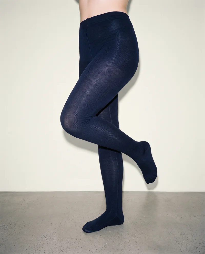 Navy wool tights shown on a female model’s lower body from a front three-quarter angle, with one knee forward. An alabaster studio-wall backdrop frames the composition.