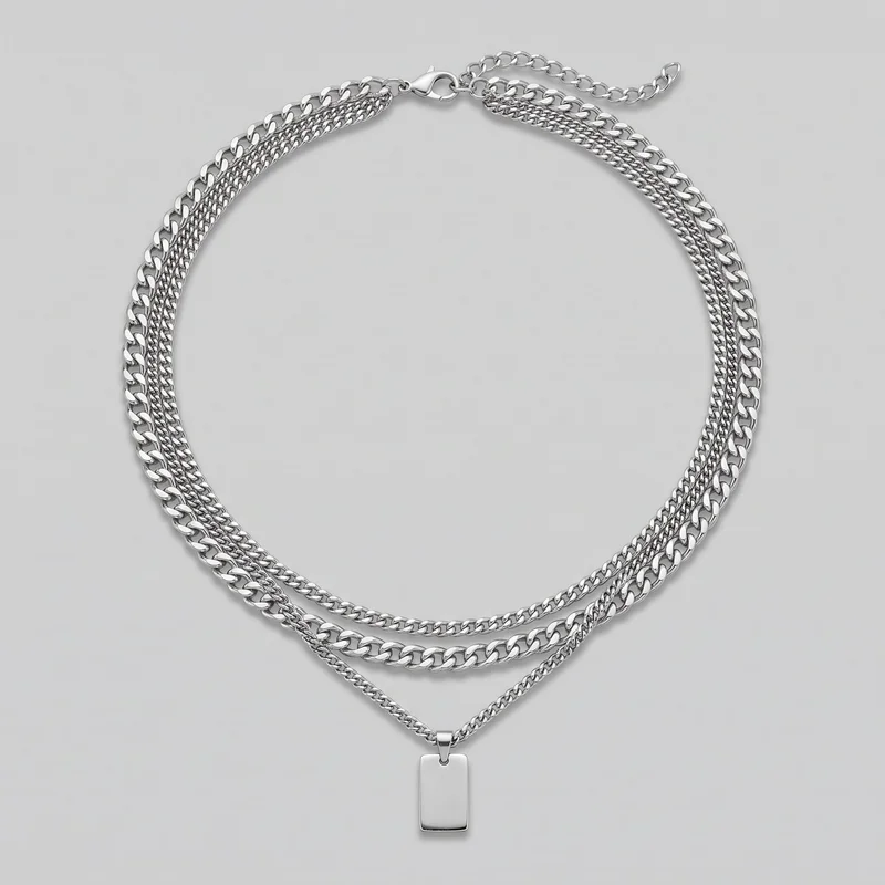 A complete men's silver layered necklace is laid flat in a loop. Multiple graduated curb-link chains frame additional curb chains through the center, with a lobster-claw clasp, extender chain, and plain vertical rectangular tag pendant visible.