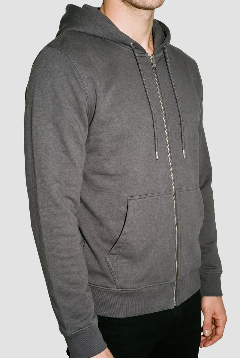 A men's charcoal cotton zip-up hoodie is shown on a male model in an angled front torso view, with the arms relaxed. The garment is presented against a flat Fog Grey backdrop.
