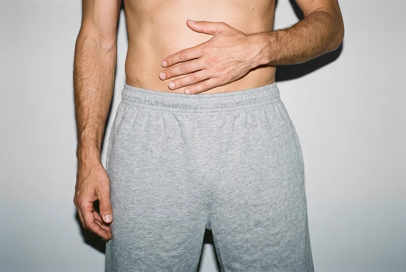 Men's grey jersey shorts shown from the front in a waistline crop, with one hand resting on the waist. A pearl grey studio-wall backdrop is visible.