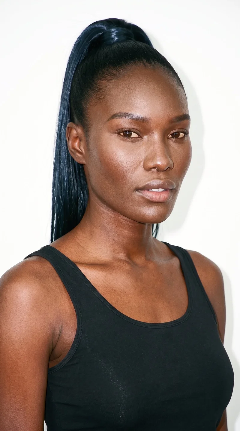 This 31-year-old African woman has dark-brown skin with a red-copper undertone. Her square face has a broad forehead and a strong, defined jaw, creating an angular, structured appearance. She has thick eyebrows, brown eyes, and medium lips. Her deep black hair is pulled into a high ponytail on the crown of her head.