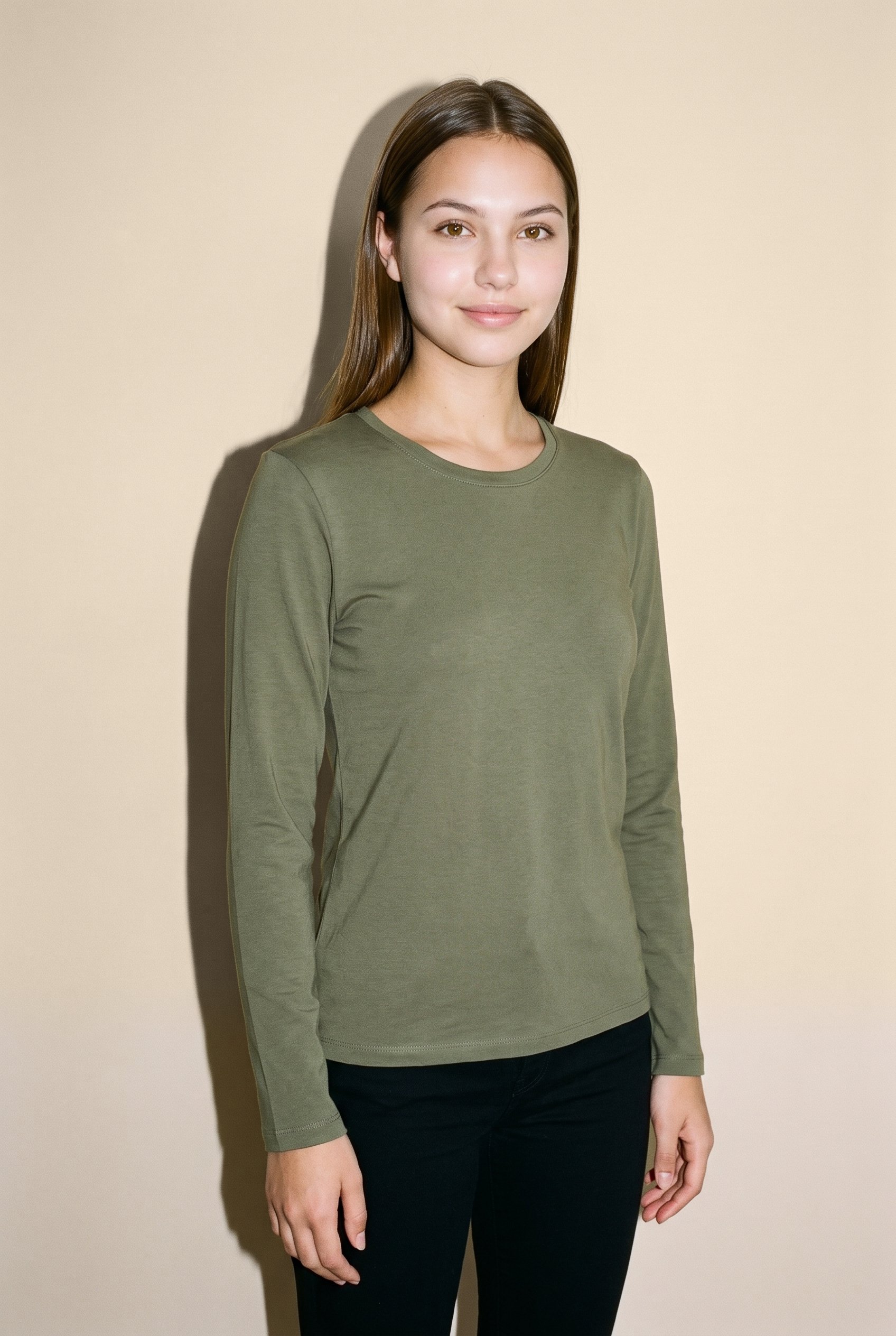 A 22-year-old Hispanic woman is shown from the upper body in an angled front view, wearing an olive cotton long-sleeve tee. Her arms are relaxed and her expression is natural, with a warm beige studio-wall backdrop.