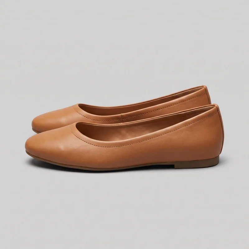 A pair of tan leather ballet flats is shown in a side-oriented standalone view. The shoes have rounded toes, shallow open toplines with stitched binding, smooth unadorned uppers, visible vamp and heel-counter seams, and thin soles with slightly raised low heels.