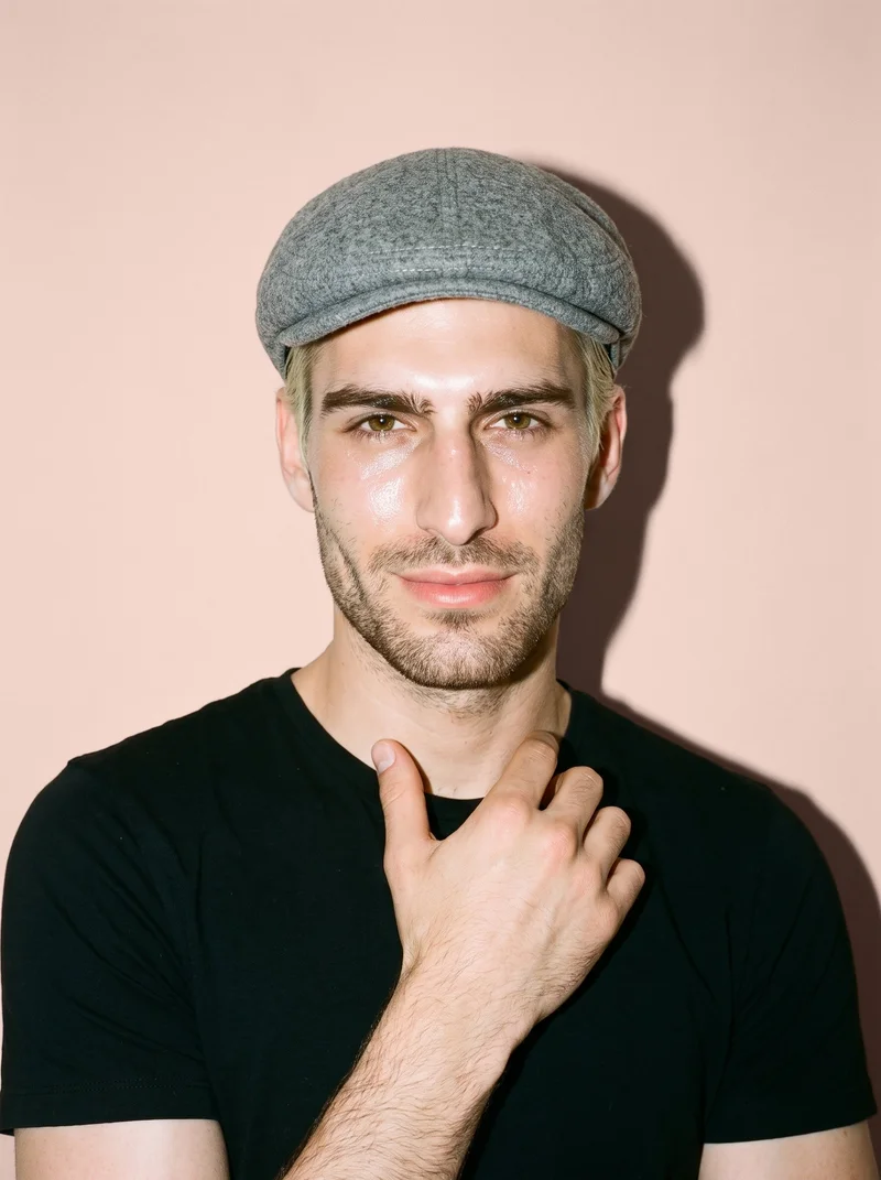 A smiling 23-year-old Caucasian man is shown in a front-facing bust view wearing a men's grey wool flat cap. One hand rests at his collarbone, with a nude blush studio-wall backdrop behind him.