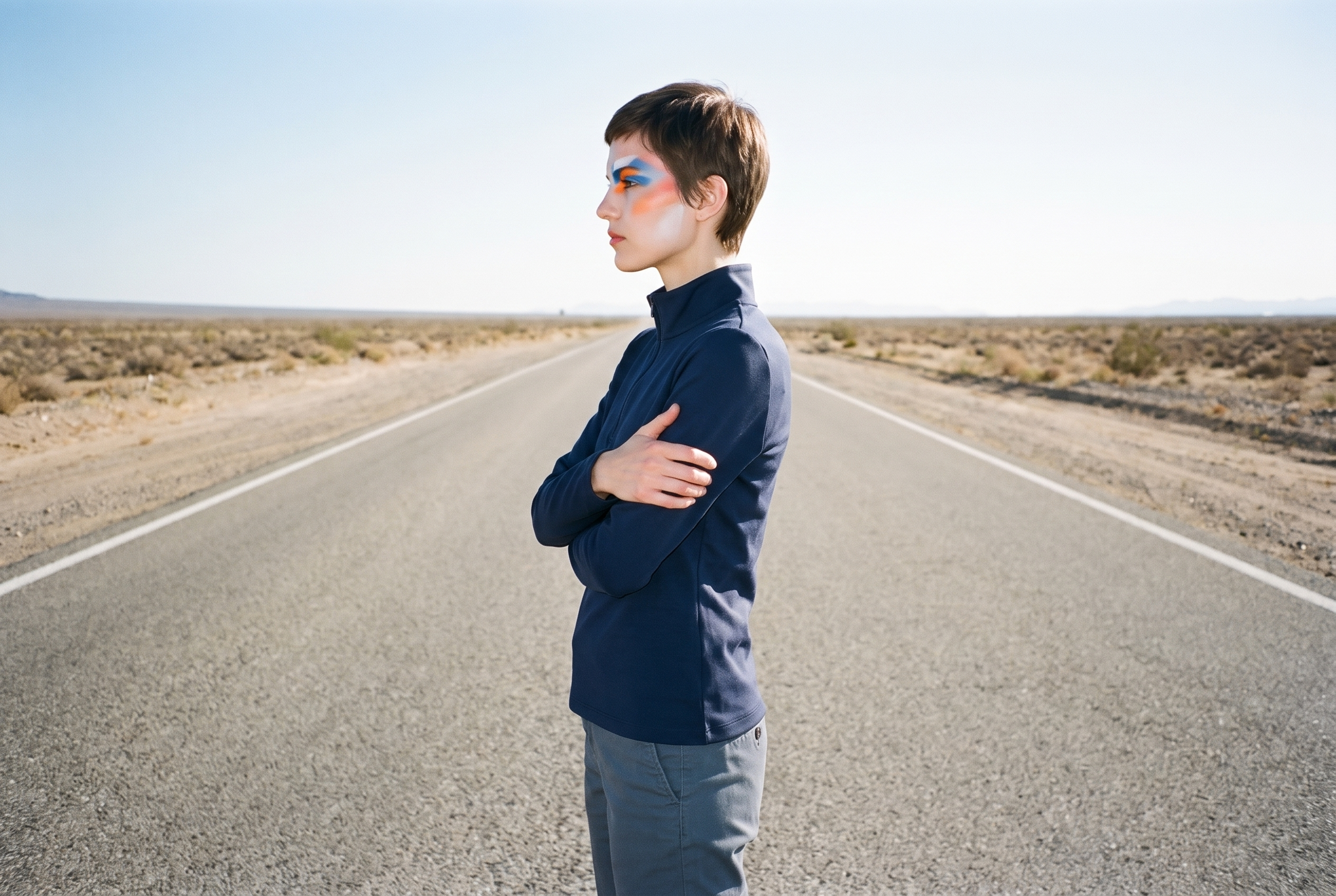 A 22-year-old Caucasian woman wears a women's navy quarter-zip in a three-quarter-length side view, holding herself in a self-hug pose with a neutral expression. A desert highway backdrop extends behind her.